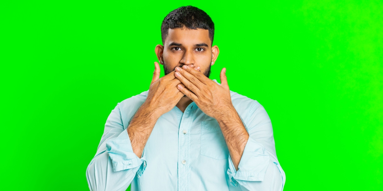An Asian man with short dark hair hold his hands up to his mouth as if silencing himself. He wears a light blue shirt. The background is green